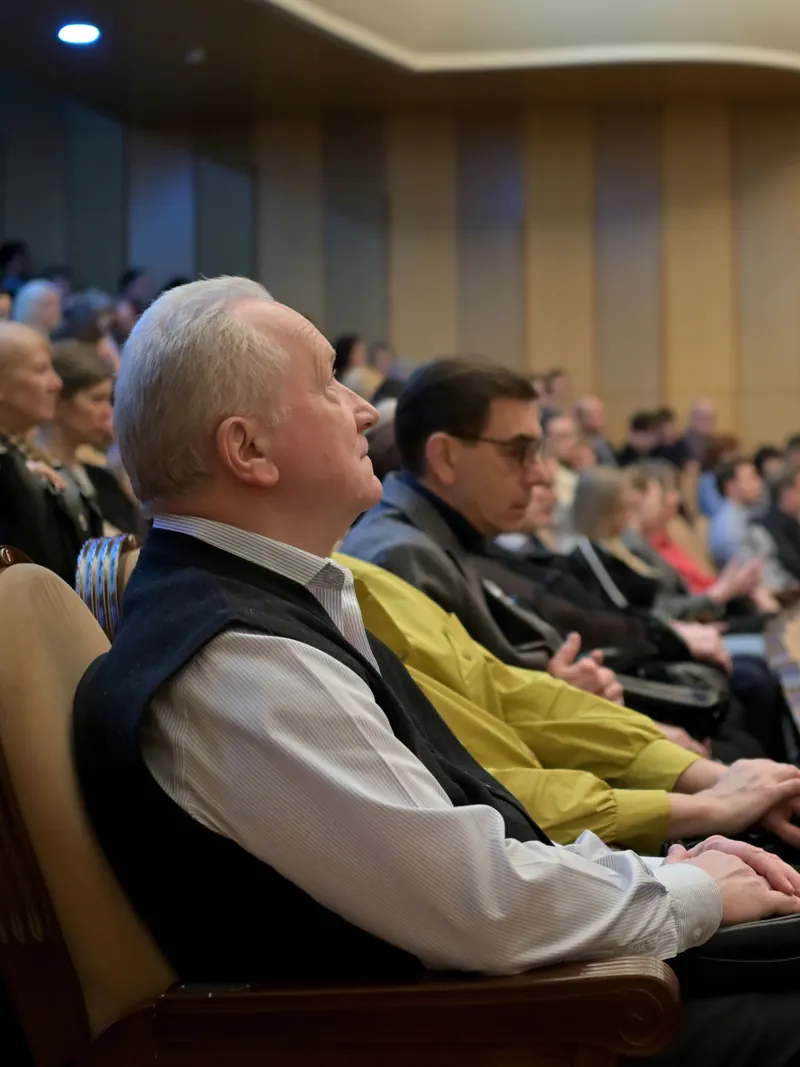 A mixed-age audience seated in a wood-panelled auditorium, seen from the side