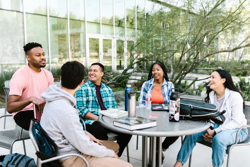 Four people around a table outside a campus building with laptops and notebooks open