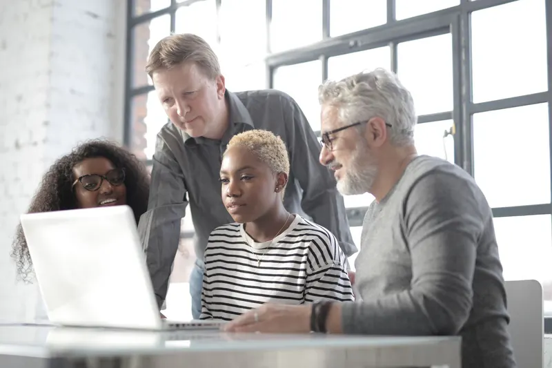 Four people of different ages gathered around one laptop, talking through what is on the screen